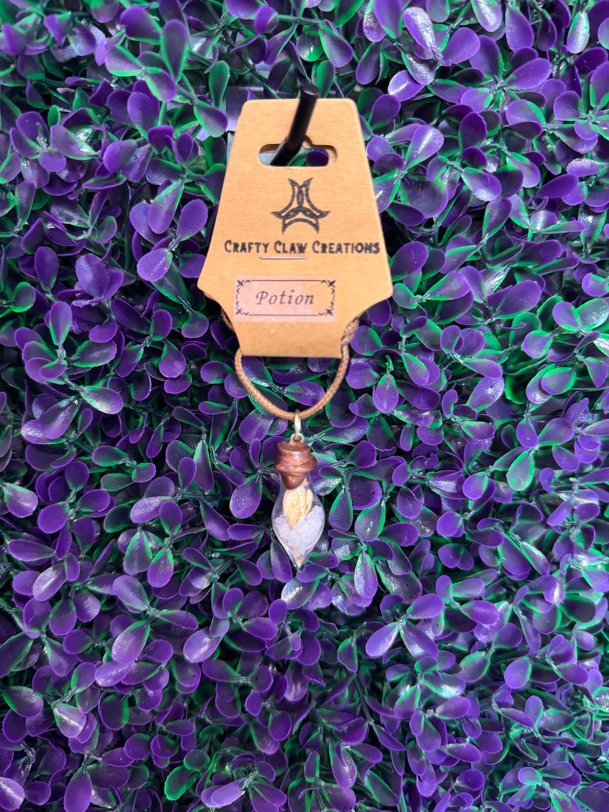 Pink Chip Potion Bottle Necklace — product photo 2. A small teardrop-shaped glass potion bottle holding pale pink chips and a golden decorative accent, finished with a copper-tone cap and tan cord.