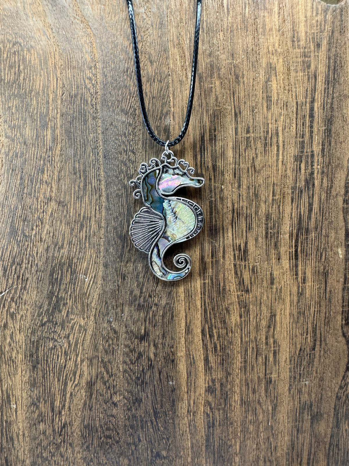 Seahorse pendant necklace with iridescent green, blue, pink, and violet shell inlay in an antiqued silver-tone metal frame, hanging from a black cord against a wood surface.