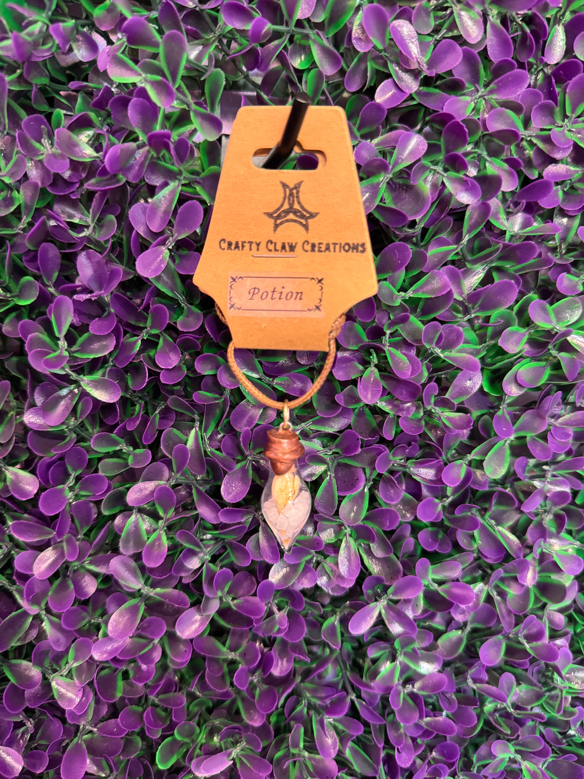 Pink Chip Potion Bottle Necklace — product photo 1. A small teardrop-shaped glass potion bottle holding pale pink chips and a golden decorative accent, finished with a copper-tone cap and tan cord.
