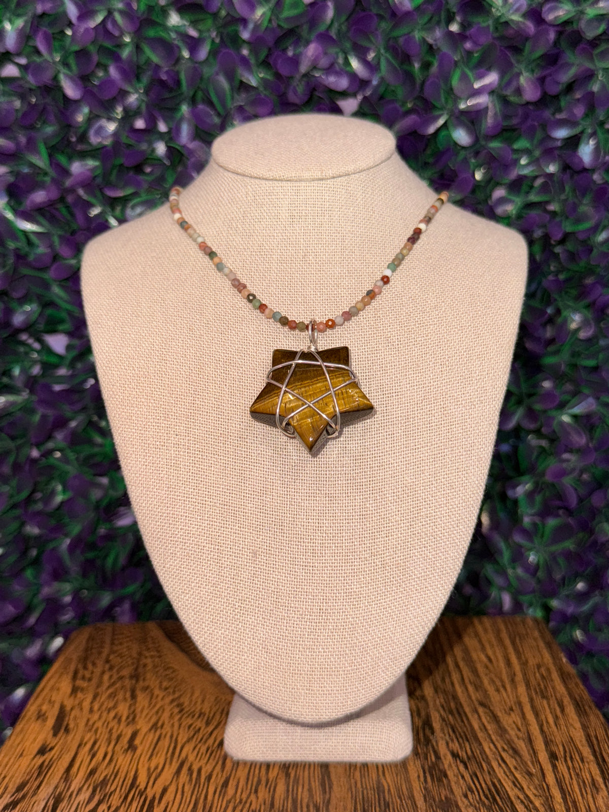 Star-shaped tiger's eye pendant wrapped in silver-tone wire on a multicolor bead chain, displayed on a linen bust.